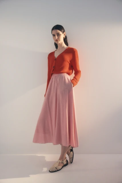Full-body shot of a young woman posing in a studio wearing a red cardigan, pink skirt, and beige sandals, with soft lighting.