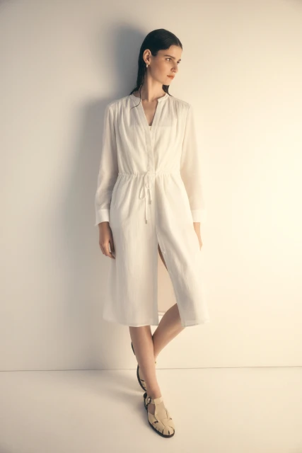 A slender adult woman with long dark hair poses in a flowing, long-sleeved white dress, set against a neutral wall and flooring backdrop.