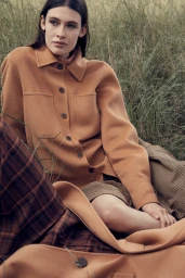 Woman in camel wool shirt jacket sitting amongst tall grass outdoors with plaid garments visible in the foreground