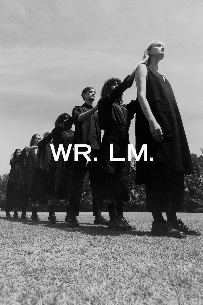 Monochrome image of a line of people standing in a row outdoors on grass, wearing dark apparel, directed by Toby Rupert
