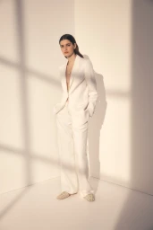 Model wearing a white pantsuit standing against an interior corner with warm window shadow lines.