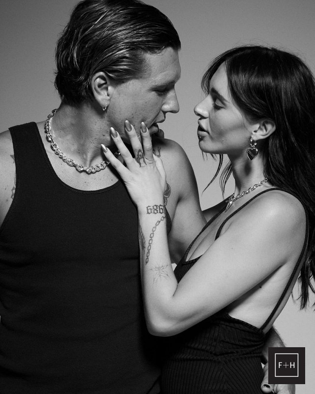 Black-and-white portrait of a couple in black tank tops and jewelry. The woman has her hand on the man's face, creating an intimate mood.