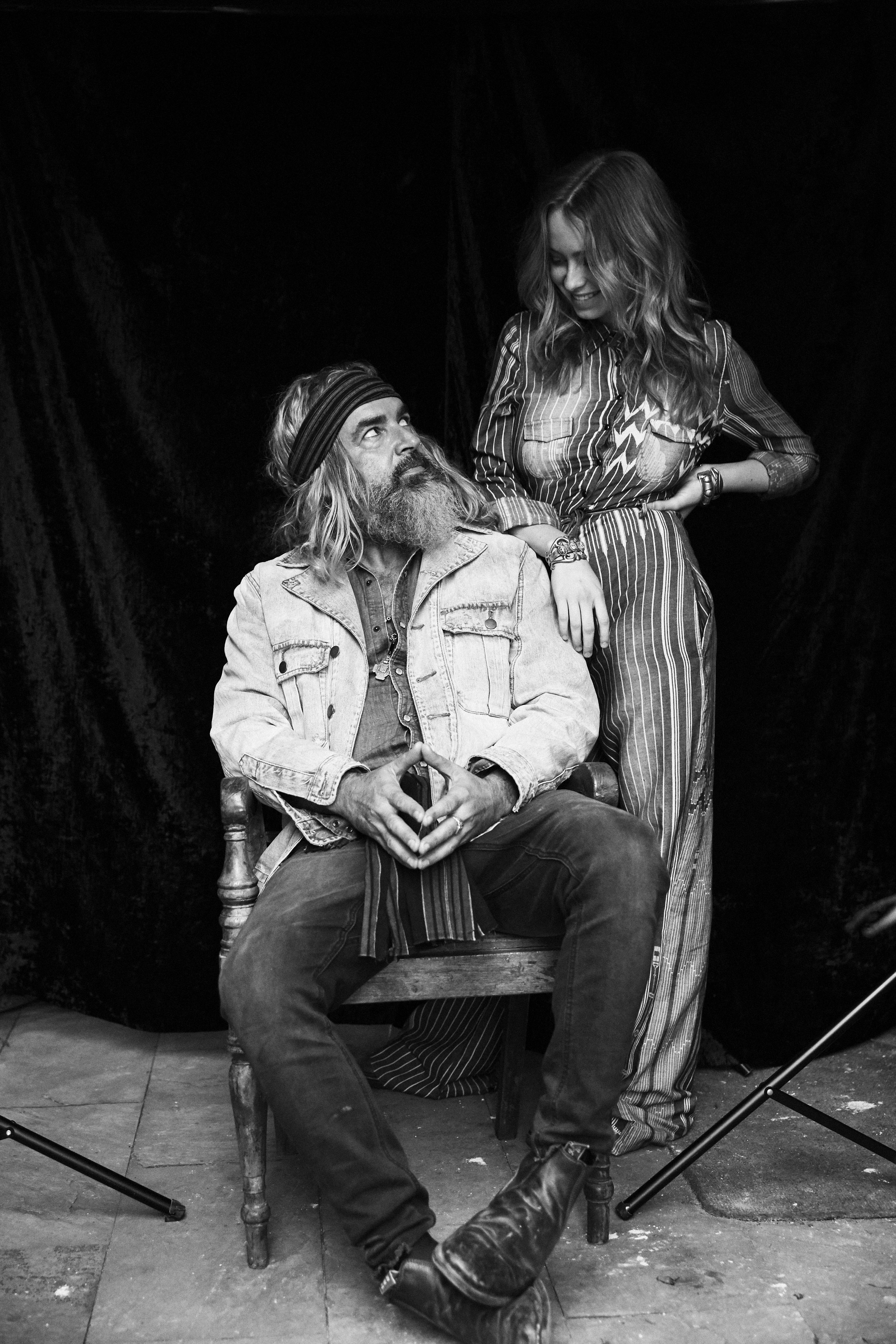 Black-and-white portrait of a long-bearded man sitting on a wooden chair looking at a young woman in a patterned jumpsuit.