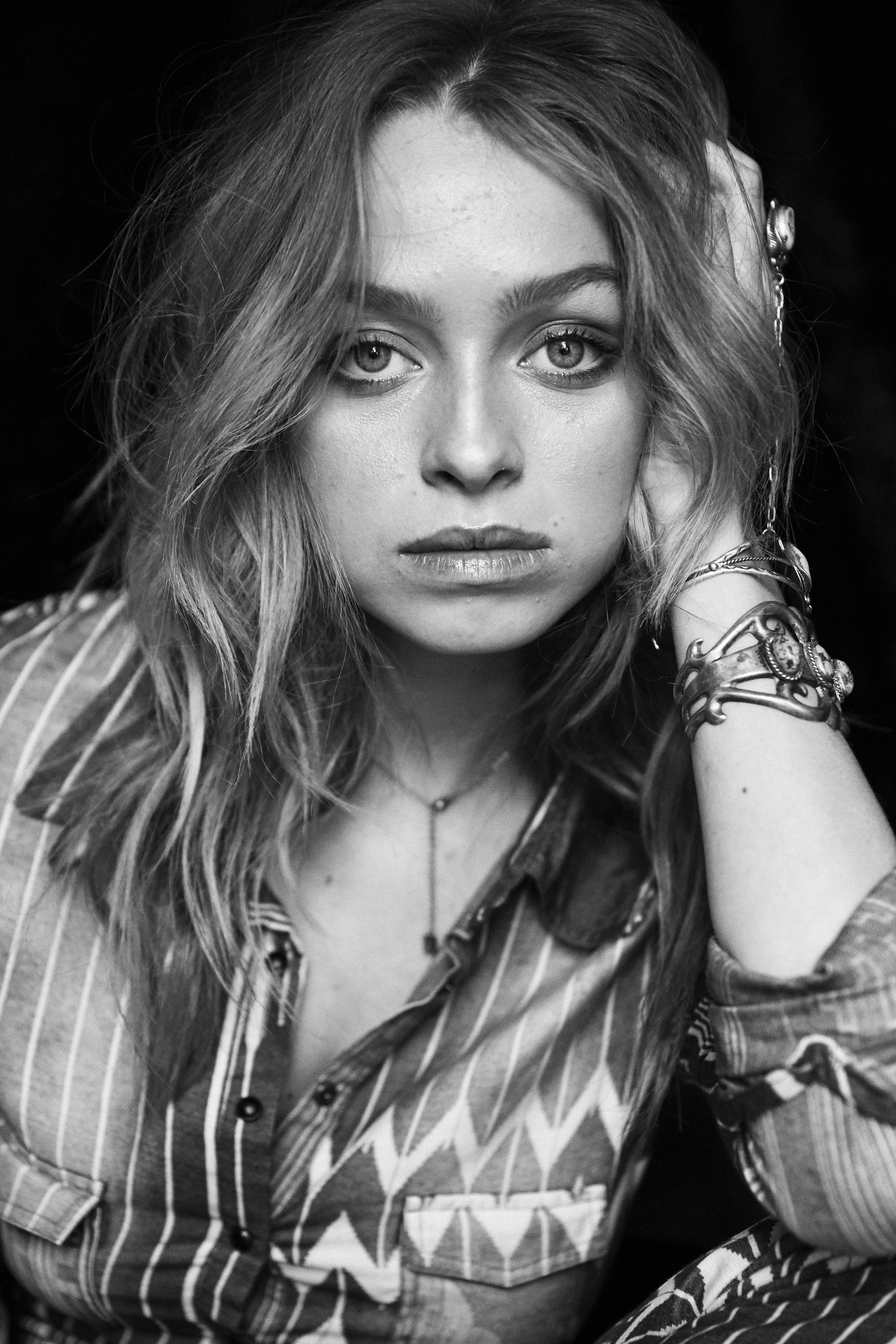 Black and white portrait of a young adult woman with wavy hair, wearing a patterned shirt, against a dark background in eye-level shot.