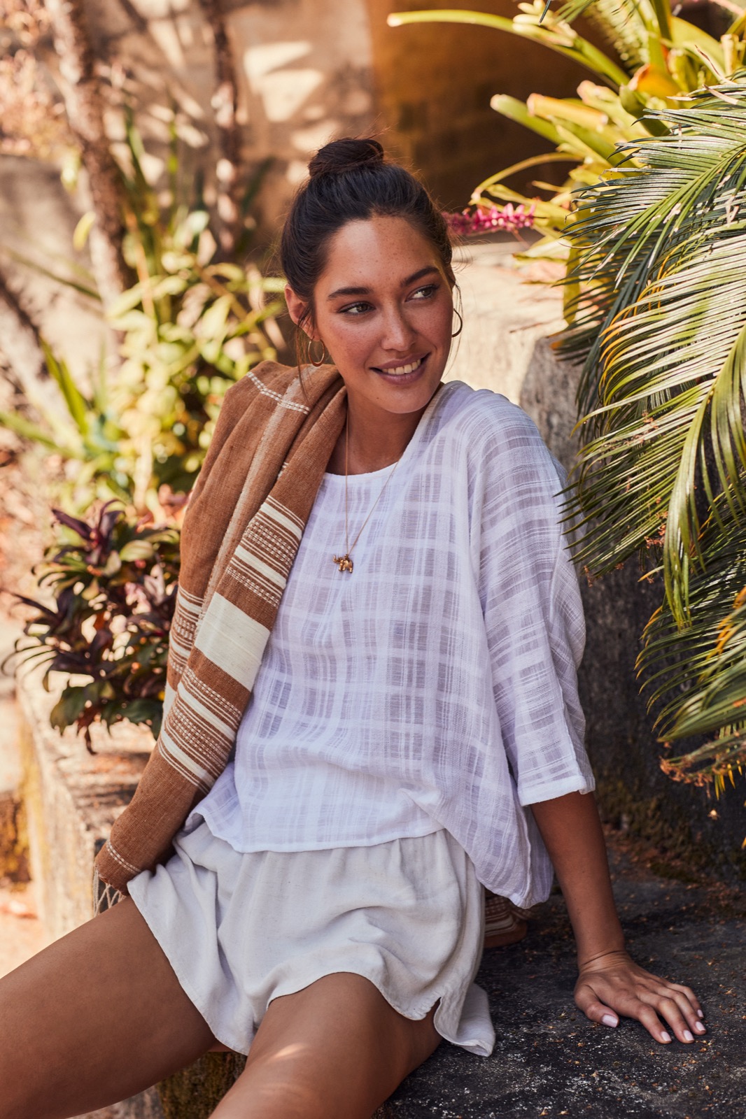 Woman in white patterned top holding a brown striped towel outdoors with lush tropical foliage background.
