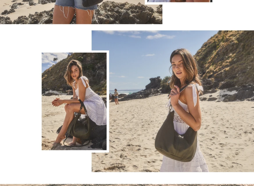 Brunette woman in white dress with olive-green bag on a sandy beach near ocean and rocks under blue sky on a sunny day.