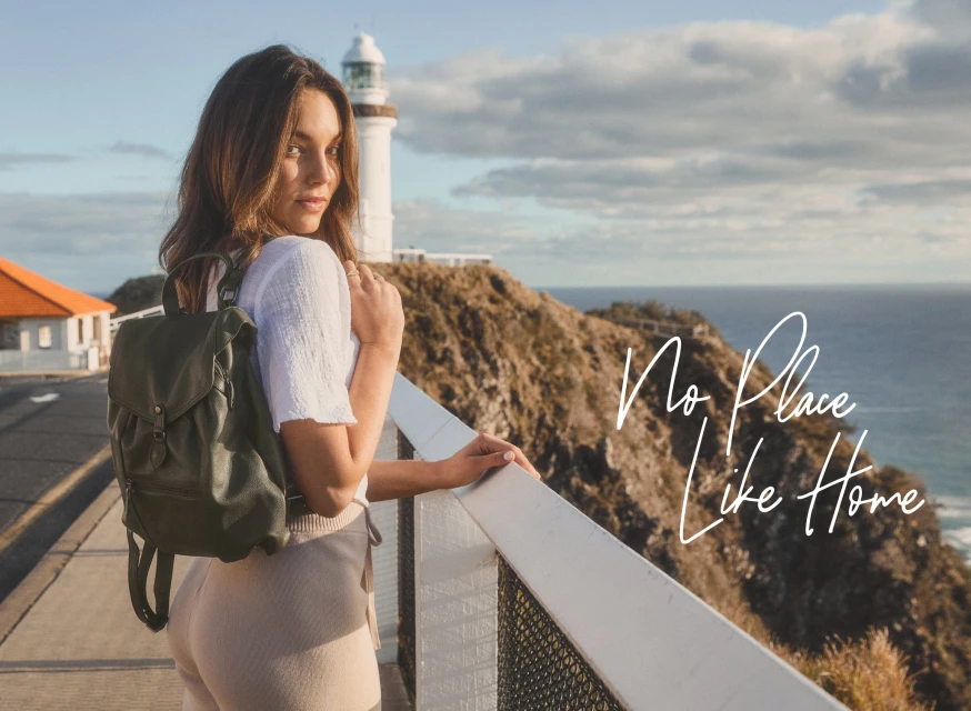 Adult woman wearing a backpack stands on a walkway overlooking a cliffside and ocean with a lighthouse in the background during daytime.