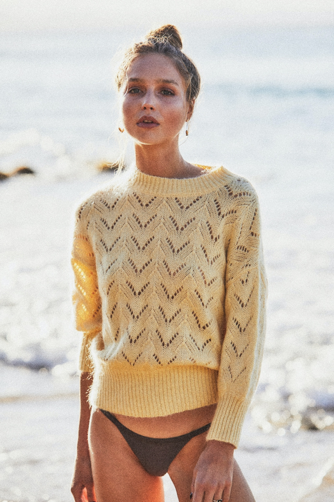 Young woman in knit sweater poses at beach on sunny day. Model with hair up, delicate jewelry, and partial view of ocean waves.