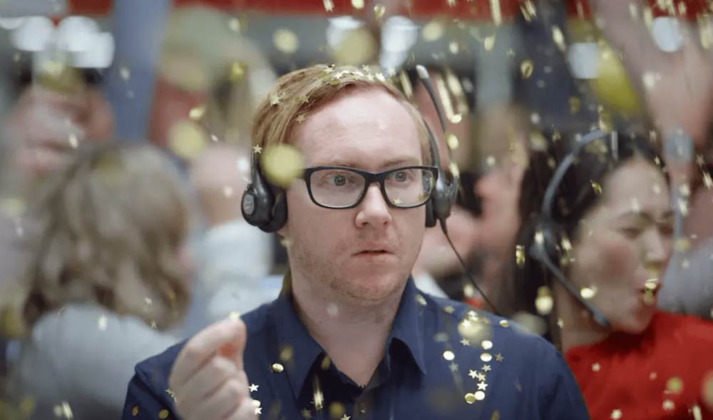 Man with headset surrounded by confetti in office with coworkers celebrating success, wearing a blue shirt and glasses indoors during daytime.