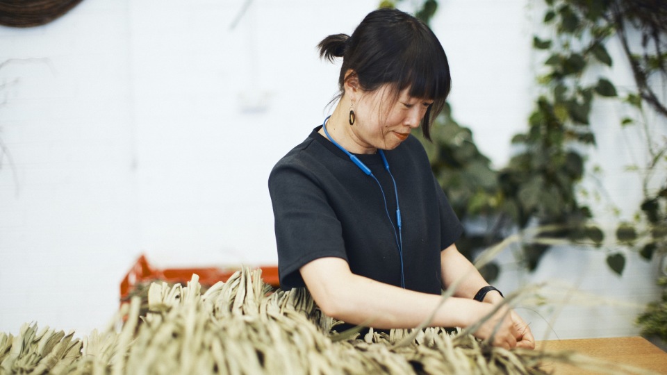 Adult woman with short dark hair in a black shirt works with dried floral elements at a table in a brightly lit indoor space.