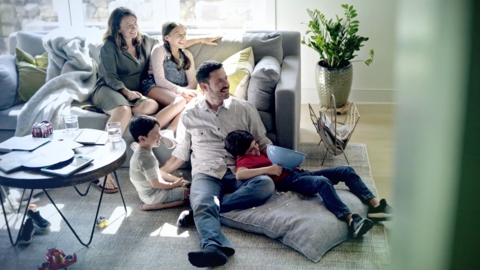 Family of six relaxing together on a couch indoors, with children sitting on a floor cushion and a potted plant in the background.