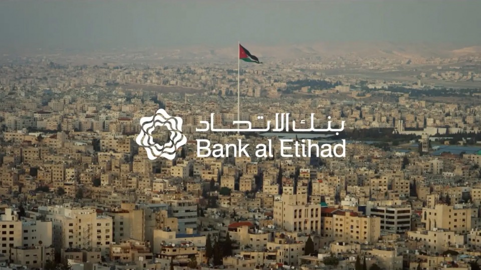 Cityscape aerial view of densely packed buildings with a waving Jordanian flag and the Bank al Etihad logo. Urban environment during daylight.