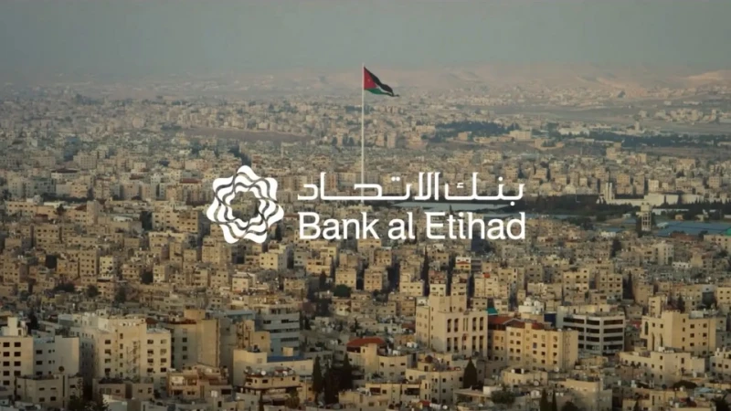Cityscape aerial view of densely packed buildings with a waving Jordanian flag and the Bank al Etihad logo. Urban environment during daylight.