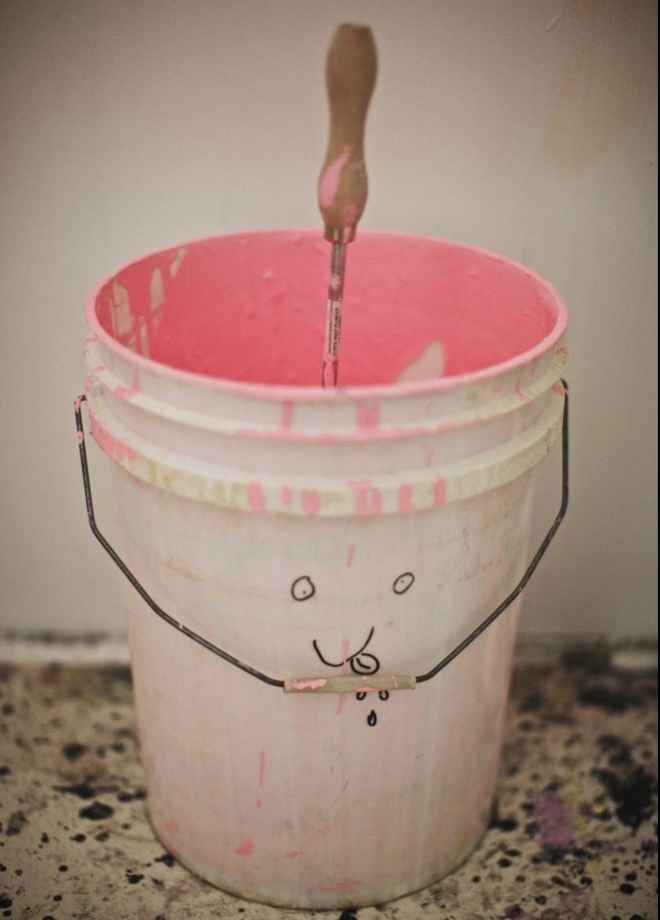 Pink paint in a bucket with a paint stirrer. A cartoon face is drawn on the bucket with pink paint splatters on the floor.