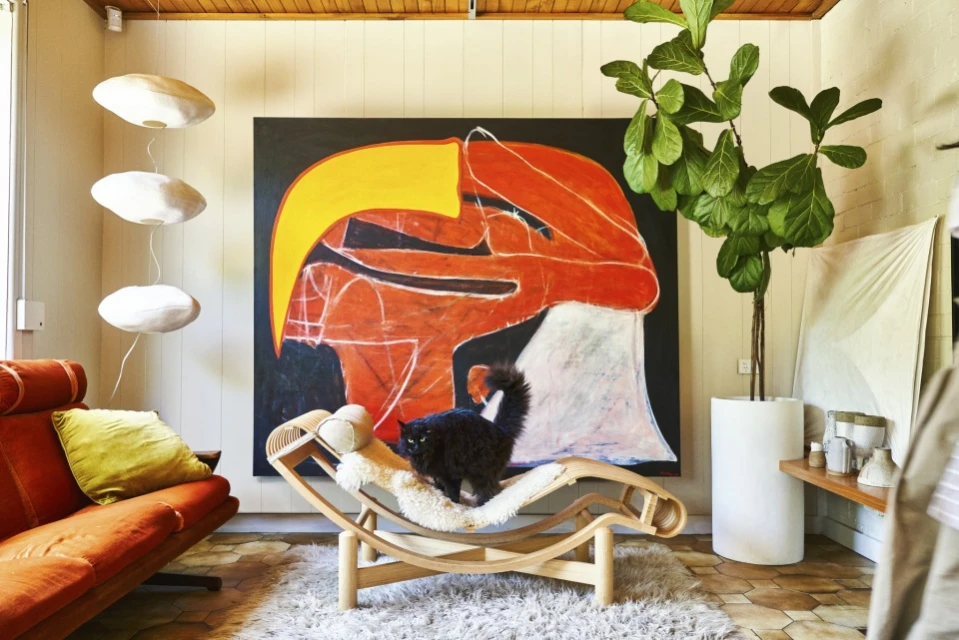 A black cat sits on a wooden lounge chair with a sheepskin cover inside a home interior, with a large painting on the wall.