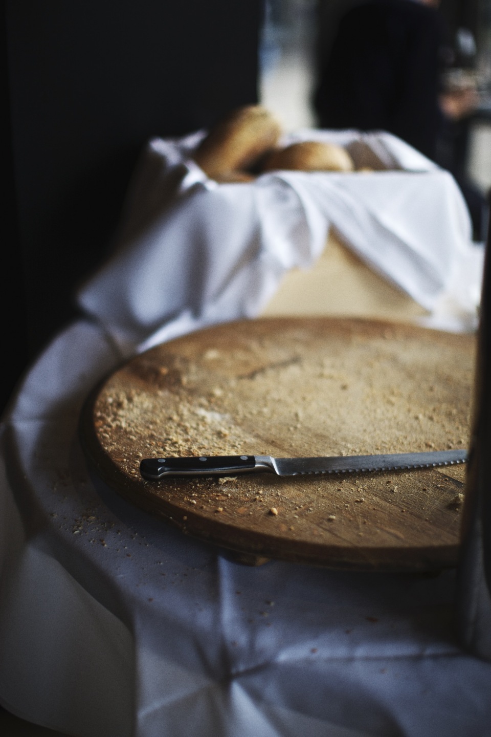 Close-up of a serrated bread knife on a round wooden cutting board covered in crumbs, with bread in a cloth-lined basket.