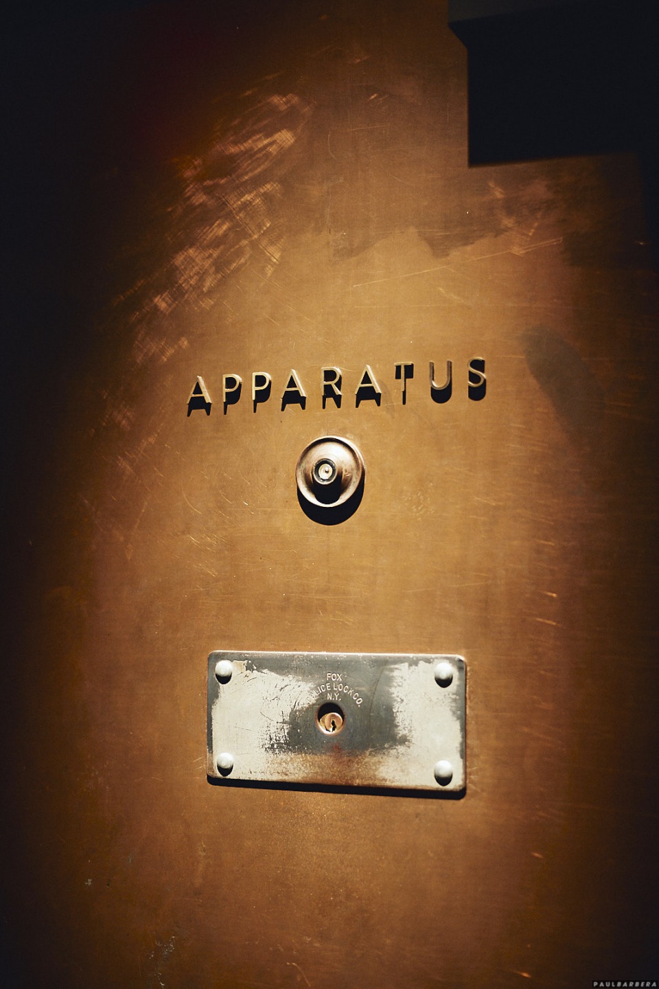 Close-up of a bronze-colored metal door with the word "APPARATUS" above a doorknob and a small rectangular metal plate below.