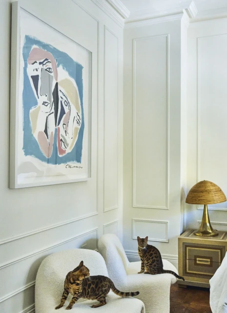 Two Bengal cats sit on white chairs in a minimalist interior featuring modern art and a woven table lamp in a well-lit room.