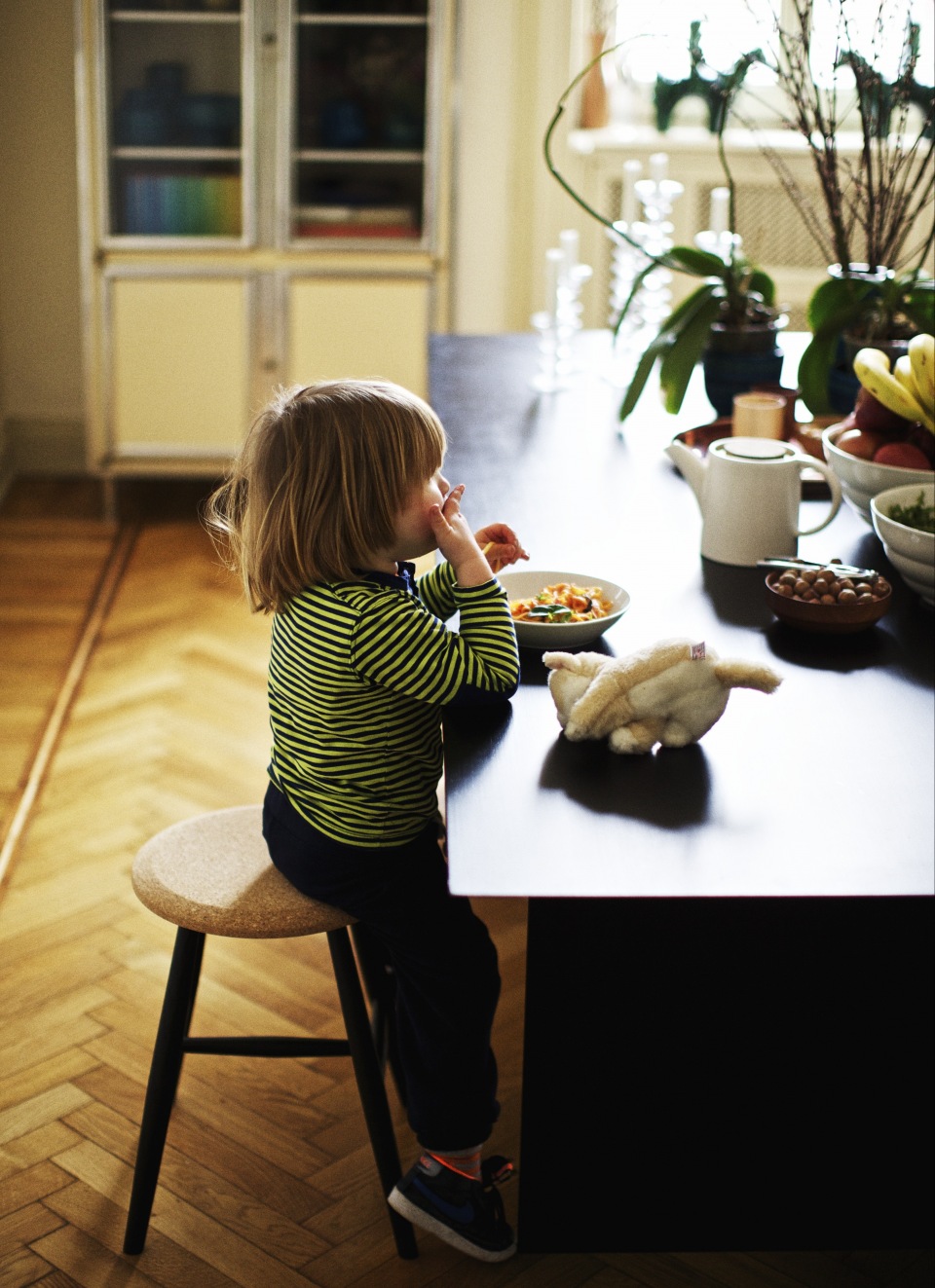 A young child with long blond hair sits on a stool at a kitchen island eating from a bowl, with a stuffed animal nearby.