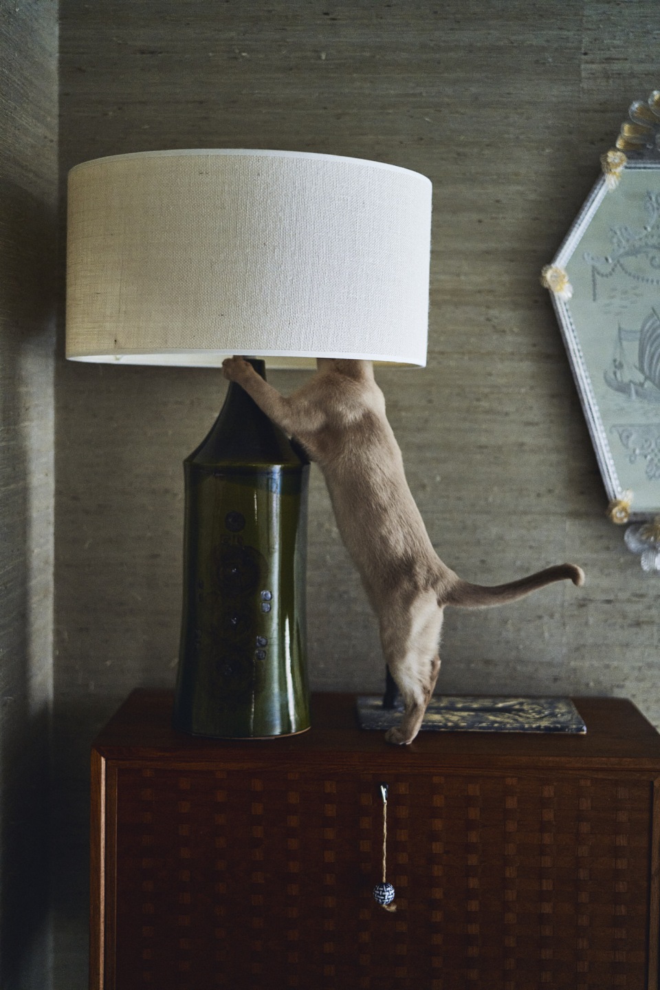 A tan cat climbs on a table lamp with a white shade and green base, situated on a wooden cabinet against a neutral, textured wall.