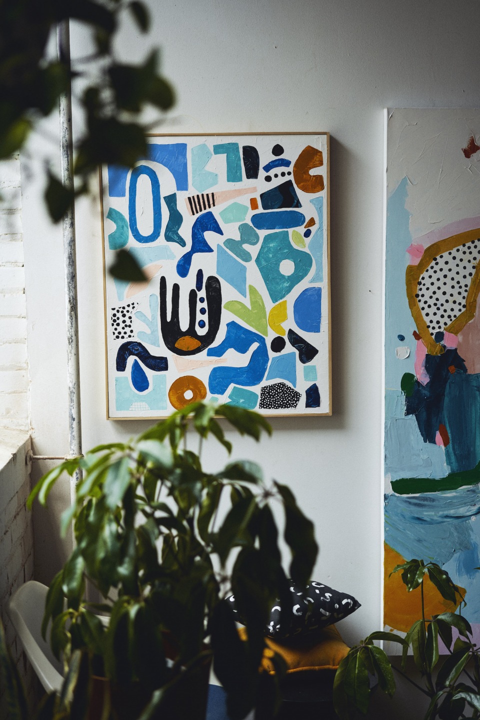 Abstract painting with blue, black, and brown shapes hanging on a wall next to another painting; leafy green plants in the foreground.