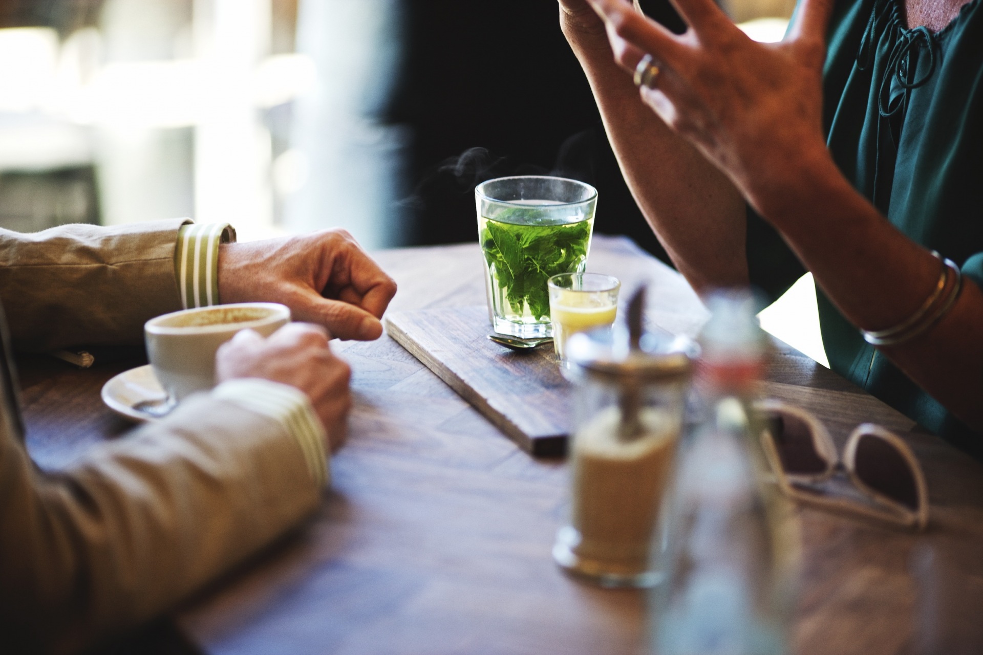 People at a cafe with glasses of tea and coffee on a wooden table. The setting is bright and airy with natural light.