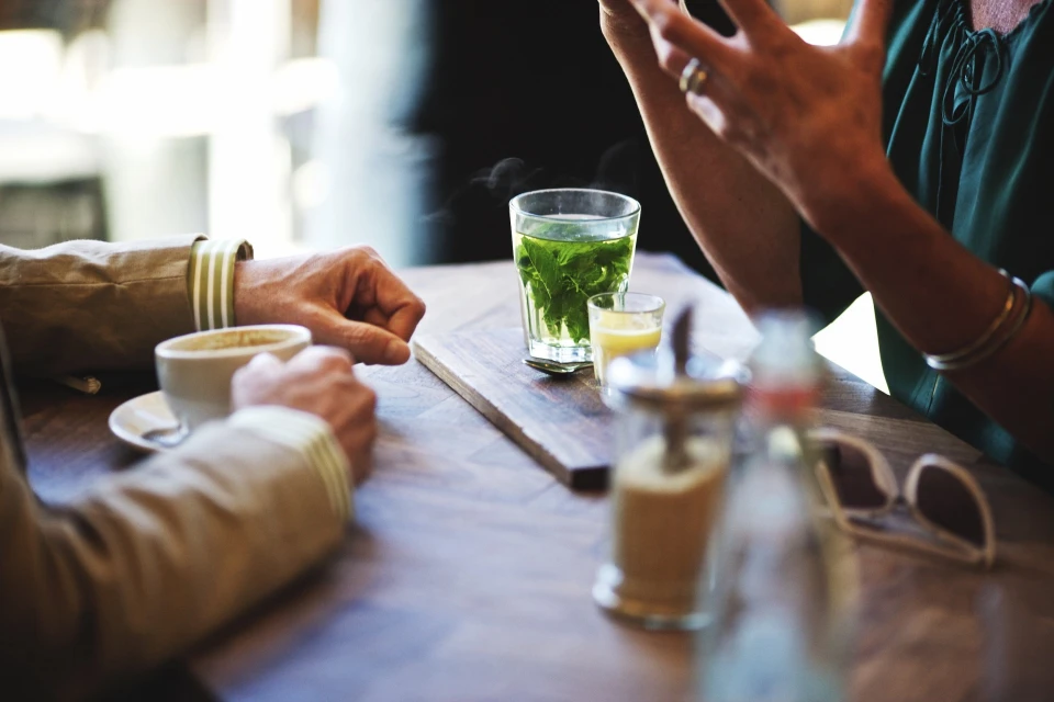 People at a cafe with glasses of tea and coffee on a wooden table. The setting is bright and airy with natural light.