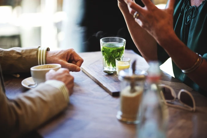 People at a cafe with glasses of tea and coffee on a wooden table. The setting is bright and airy with natural light.