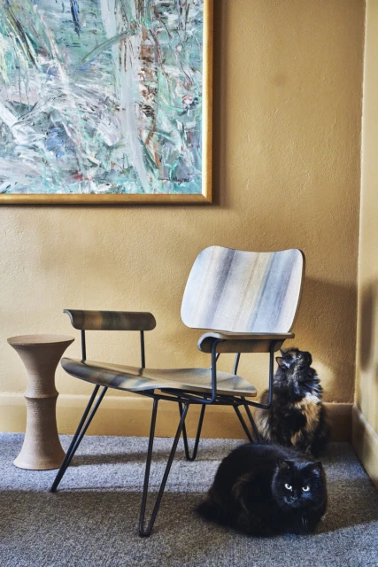 Indoor shot featuring a mid-century modern chair, an abstract painting, decorative stool, and two cats, one black and one multi-colored.