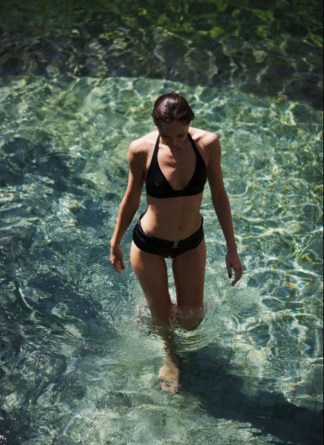 Woman in black bikini walking in clear, shallow water with visible ripples and reflections, creating a serene and natural scene.
