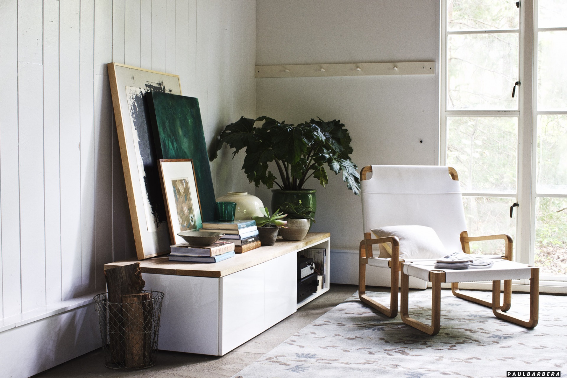 Living room interior featuring a white lounge chair, plants in pots, and decorative objects on a low cabinet in a brightly lit space.