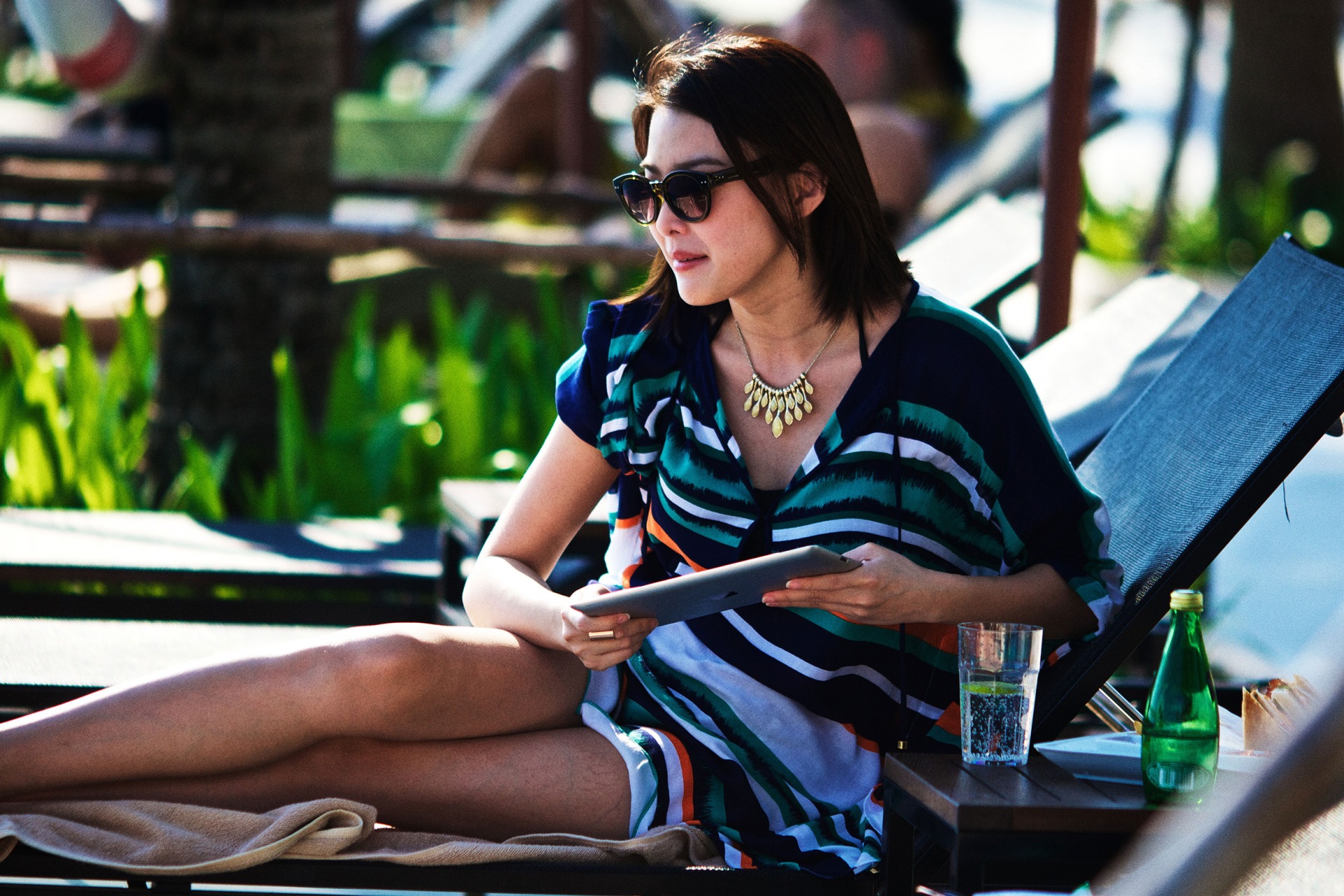 Adult woman with sunglasses relaxing poolside on a lounge chair and using a tablet with drinks in the background on a sunny day.
