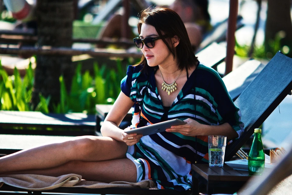 Adult woman with sunglasses relaxing poolside on a lounge chair and using a tablet with drinks in the background on a sunny day.