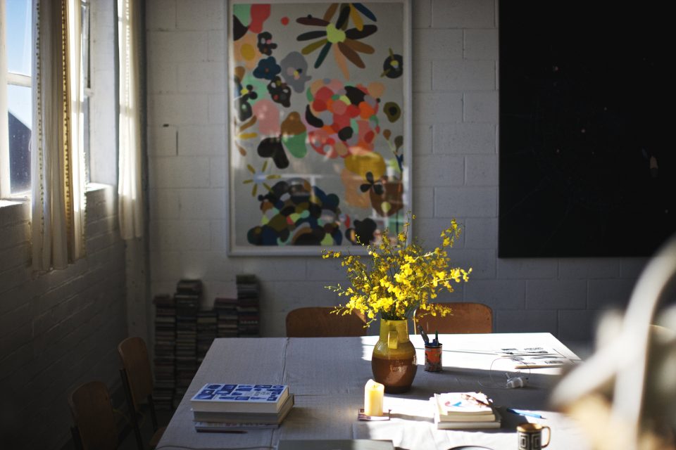 Still life interior with an abstract art print hanging above a table with a yellow flower arrangement, books, and window light.