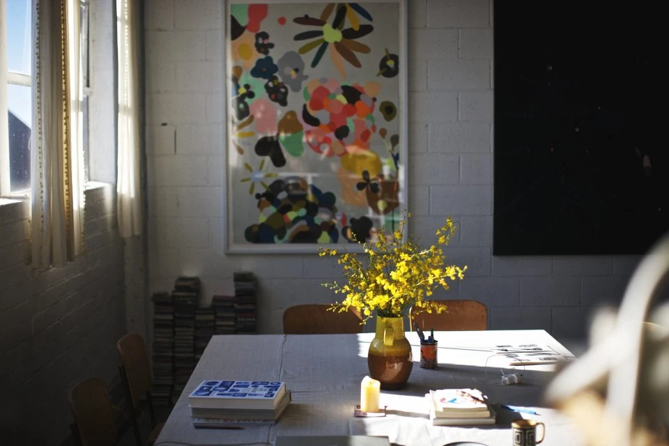 Still life interior with an abstract art print hanging above a table with a yellow flower arrangement, books, and window light.