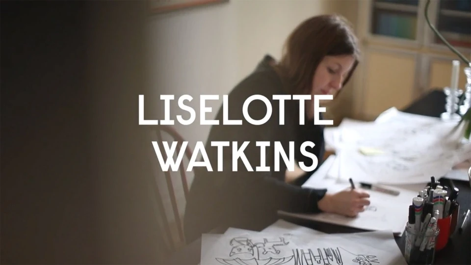 Adult woman draws on papers on a table with drawing implements; warm indoor light with a glimpse of shelves in the background.