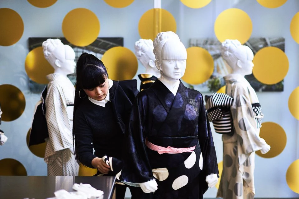 Adult female adjusts a traditional Japanese kimono on a mannequin in a studio; other mannequins stand in background with gold circles.