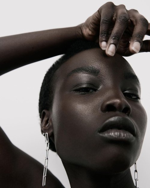 Dark-skinned woman's portrait with silver chain earrings against a clean, light background in a beauty or fashion editorial style.