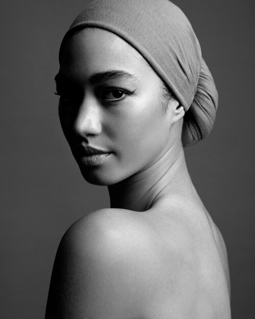 Black and white portrait of a young woman with a headscarf, looking over her shoulder with a neutral expression in a photo studio.
