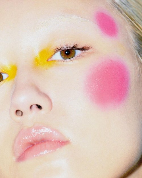 Close-up portrait of a fair-skinned model with creative yellow and pink makeup on her face, glossy lips, and soft diffused lighting.