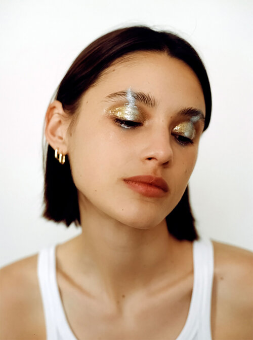 Close-up shot of a young woman with glittery gold and silver makeup on her eyes and eyebrows, posing indoors against a white background.