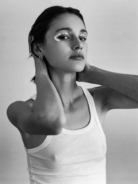 Black and white portrait of a young adult woman with silver makeup around her eyes, wearing a white ribbed tank top.