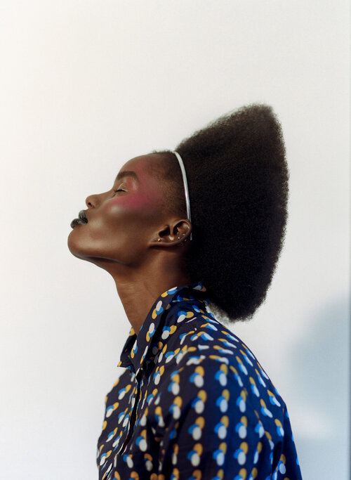Profile portrait of a dark-skinned woman with an afro, wearing a patterned shirt against a white background, emphasizing natural beauty.