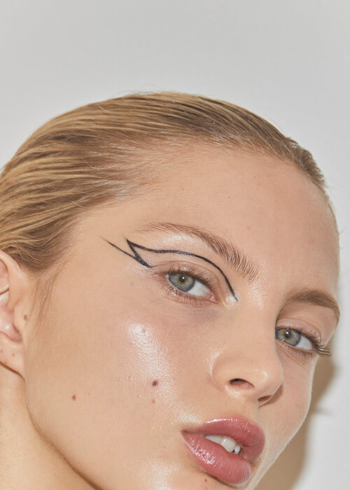Close-up portrait of a blonde woman with dramatic black eyeliner, glossy skin, and a fresh makeup look against a plain white backdrop.