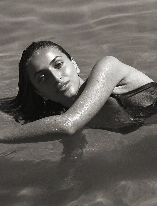 Monochrome portrait of a woman with dark hair partially submerged in water, resting her arms on the surface in an outdoor setting.