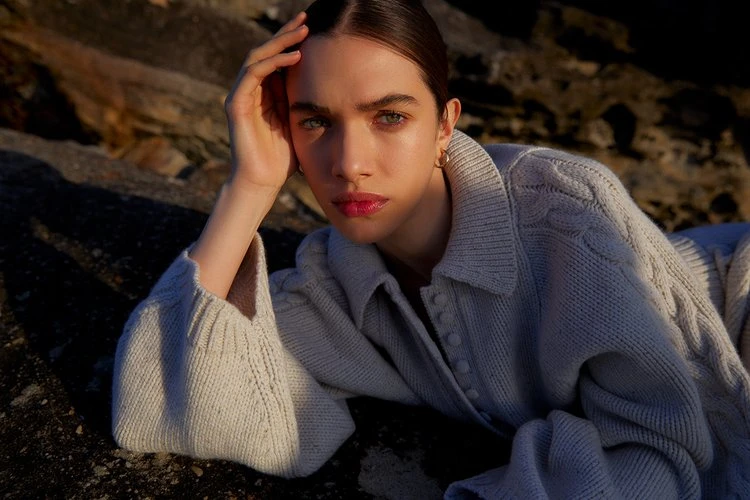 A young adult woman with green eyes and pink lipstick poses in a cable knit cardigan sweater on a rocky surface with golden lighting.
