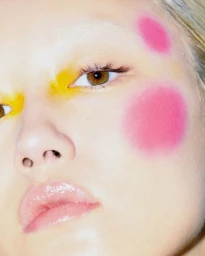Close-up portrait of a model featuring vibrant yellow eye makeup and bold pink circles on cheek and temple, makeup by Katie Angus.