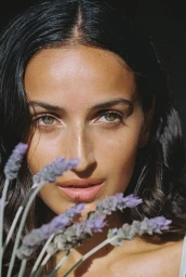 Close-up portrait of a woman with dark wavy hair and green eyes, holding sprigs of purple lavender in the foreground.