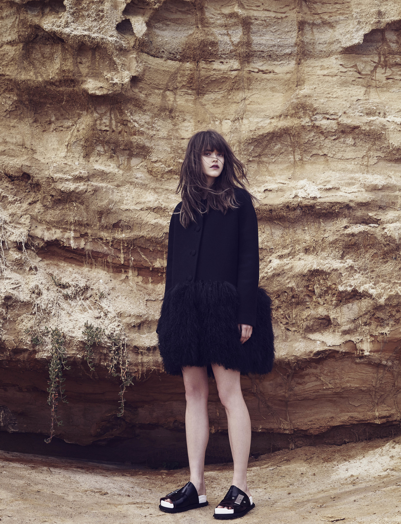 Woman in a dark coat with a shaggy trim standing in front of a layered tan rock wall with dark hair and black flats.