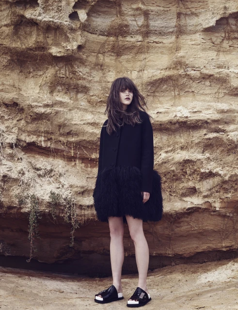 Woman in a dark coat with a shaggy trim standing in front of a layered tan rock wall with dark hair and black flats.
